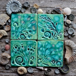 Puede incluir: Cuatro baldosas cuadradas de cerámica turquesa con diseños botánicos y conchas marinas en relieve. Las baldosas están rodeadas de varias conchas marinas y fósiles sobre una superficie de madera envejecida. Las baldosas presentan patrones detallados de helechos, espirales y otras criaturas marinas.