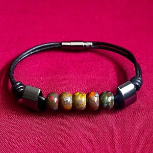 May include: Black leather bracelet with a magnetic clasp. The bracelet features six multi-coloured, patterned beads and two silver-toned, rectangular metal accents. The bracelet is displayed on a red background.