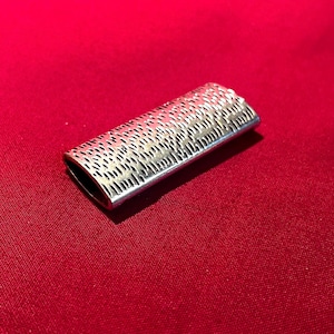 May include: A silver-colored, rectangular metal object with a textured, perforated design. The object has rounded ends and appears to be a decorative or functional item. The background is a solid red color.