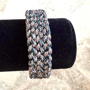 May include: A wide, braided bracelet in a mix of colours including grey, teal, and brown. The bracelet is made of a thick, woven material, and is displayed on a black velvet surface. The bracelet's design is intricate and textured.
