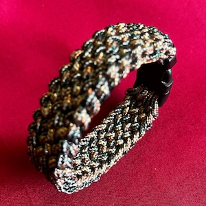 May include: A braided bracelet with a black plastic clasp. The bracelet is made of a multicolored cord in shades of black, green, brown, and gold. The bracelet is shown against a red background.