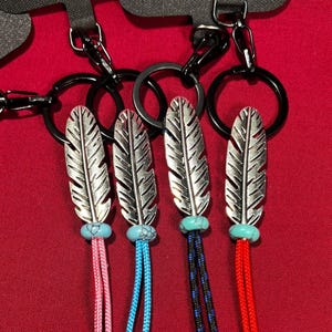 Puede incluir: Cuatro colgantes de plumas plateadas con cuentas turquesas y cordones coloridos. Los cordones son rosa, azul, negro y azul, y rojo. Cada colgante está unido a un anillo negro y una correa negra.