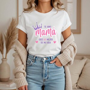 Puede incluir: Camiseta blanca con el texto "TE AMO Mamá ERES LO MEJOR DE MI VIDA" en rosa y morado. El diseño incluye una corona y corazones. La camiseta se combina con vaqueros azules y una rebeca beige.