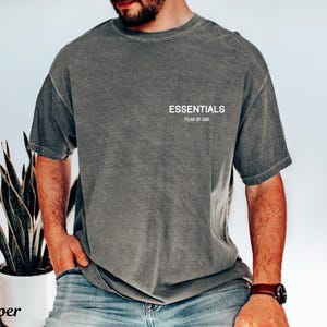 May include: Dark grey t-shirt with the word "ESSENTIALS" printed in white on the chest. The text "FEAR OF GOD" is printed below the word "ESSENTIALS". The shirt has short sleeves and a crew neck.