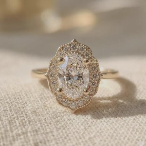 May include: A gold ring featuring a central oval-cut clear gemstone surrounded by a scalloped halo of small round clear stones. The band is thin and polished, showcasing a detailed metal setting that frames the oval center stone with precision.