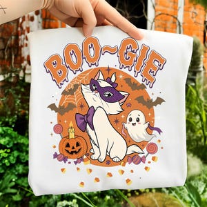 May include: A white t-shirt featuring a graphic of a white cat wearing a purple mask and bow tie, a ghost, a jack-o'-lantern, bats, and candy. The text BOO-GIE is printed in orange above the cat. The design includes a spiderweb and stars.