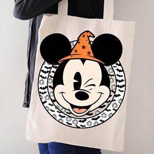 Könnte beinhalten: Eine cremefarbene Stofftasche mit einer Grafik von Mickey Mouse, die einen orangefarbenen Hexenhut mit schwarzen Sternen trägt. Die Figur ist in einem kreisförmigen Rahmen zentriert, der mit kleinen schwarzen Fledermäusen und weißen Kürbissen verziert ist.