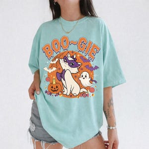 Op de afbeelding: Een licht mintgroen t-shirt met een grafische print met de oranje tekst BOO-GIE. Het ontwerp bevat een witte kat met een paars masker en strikje, een klein spookje, een pompoen, vleermuizen, een spinnenweb en kleurrijke snoepjes.