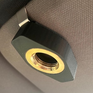 May include: A black, geometric-shaped car accessory with a gold-colored interior ring. The accessory is attached to a gray fabric surface, possibly a car's interior. The design is modern and minimalist.