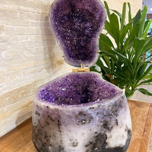 May include: A large amethyst geode sculpture with a deep purple crystal interior. The geode is mounted on a wooden base with gold-colored supports. The exterior is a mix of white, gray, and black.