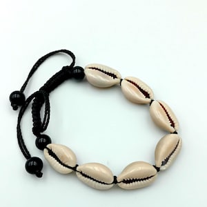 Peut inclure: Un bracelet composé de coquillages beiges, enfilés sur un cordon noir. Le bracelet a une fermeture coulissante avec deux perles noires. Les coquillages ont une texture naturelle. Le bracelet mesure environ 18 cm de long.