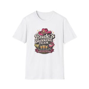 Può includere: T-shirt bianco con un design per un addio al nubilato. Il design include un cappello da cowboy rosa, il testo "Bride's Drinking Team" e immagini di cocktail e shot. È presente anche il testo "Buy us a shot, we're tying the knot".