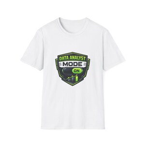 Può includere: T-shirt bianco con grafica verde e nera con la scritta "DATA ANALYST MODE ON". La grafica è a forma di scudo con un grafico a barre e un interruttore. La maglietta ha maniche corte e girocollo.