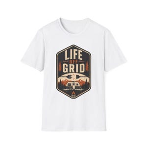 Può includere: T-shirt bianca con un design grafico. Il design presenta il testo "LIFE OFF GRID", un camper, montagne, alberi e un falò. La grafica è nei toni del marrone, arancione e nero.