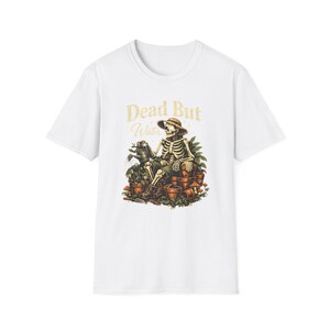 Può includere: T-shirt bianca con una grafica umoristica di uno scheletro con un cappello di paglia che innaffia le piante. Il testo "Dead But Water" è sopra l'illustrazione. Il design include toni marroni e verdi.