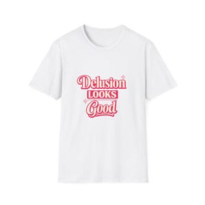 Può includere: T-shirt bianca con la scritta "Delusion Looks Good" in un font retrò e corsivo. Il testo è di colore rosa-rossastro, con stelle. Un capo casual per tutti i giorni.