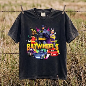 Pode incluir: Uma t-shirt preta de manga curta com uma impressão gráfica do Batman, Robin e Batgirl de pé sobre três veículos animados. O texto BATWHEELS está impresso em letras amarelas grandes no centro do design gráfico.