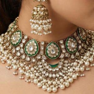 Puede incluir: Un conjunto de collar y pendientes dorados ornamentados. El collar presenta detalles de esmalte verde, piedras claras en forma de lágrima y perlas blancas colgantes. Los pendientes a juego tienen un diseño similar con perlas colgantes.