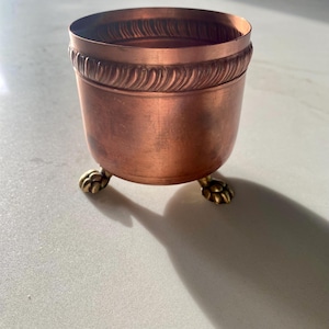 Puede incluir: Un cuenco de metal color cobre con un cuerpo cilíndrico liso y una textura decorativa similar a una cuerda alrededor del borde superior. El cuenco se apoya sobre tres pequeños pies de metal dorado con forma de patas de animal con garras.