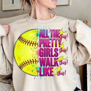 May include: A cream-colored sweatshirt featuring a large softball graphic with the text "ALL THE PRETTY GIRLS WALK LIKE THIS!". The softball is yellow with red stitching, and the text is pink and purple.