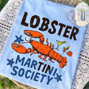 Pu&ograve; includere: T-shirt azzurro con grafica di un'aragosta arancione, un bicchiere da martini e la scritta "LOBSTER MARTINI SOCIETY" in marrone e blu. Il design include stelle marine e una conchiglia.