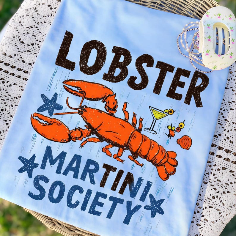 Pu&ograve; includere: T-shirt azzurro con grafica di un'aragosta arancione, un bicchiere da martini e la scritta "LOBSTER MARTINI SOCIETY" in marrone e blu. Il design include stelle marine e una conchiglia.