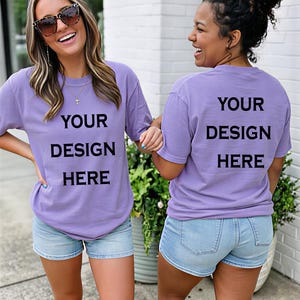 Op de afbeelding: Twee vrouwen dragen lavendelkleurige T-shirts en spijkerbroekshorts. De shirts hebben de tekst "YOUR DESIGN HERE" in het zwart gedrukt. Eén vrouw draagt een zonnebril. De achtergrond omvat groen en een witte bakstenen muur.