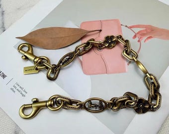 May include: A brass-toned metal chain strap featuring oval links, two lobster claw clasps, and decorative floral and horse-and-carriage motifs. A small rectangular tag with the word COACH is attached to one end of the chain, which has a vintage finish.