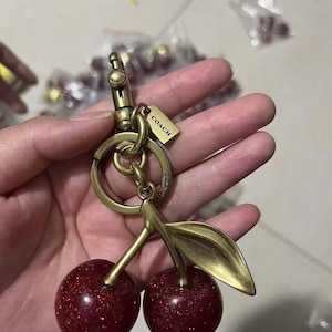 May include: A cherry-shaped keyring featuring two dark red glittery spheres attached to a brass-toned metal stem and leaf. The hardware includes a circular ring and a lobster clasp, with a small rectangular tag engraved with the word COACH.