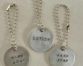May include: Three silver metal tags on ball chains. Each tag is round with stamped black lettering. The tags read "DISH SOAP", "LOTION", and "HAND SOAP". The tags are hanging against a light background.