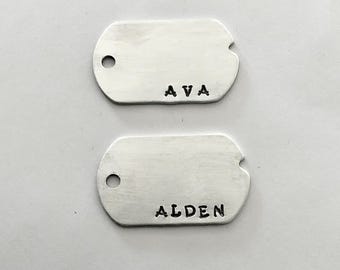 May include: Two silver dog tags with rounded edges and a hole at the top. The top tag reads "AVA" in black letters, and the bottom tag reads "ALDEN". The tags are set against a white background.