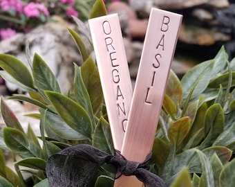 May include: Two metal plant markers, one labeled "BASIL" and the other "OREGANO", are stuck into a potted plant with green leaves.