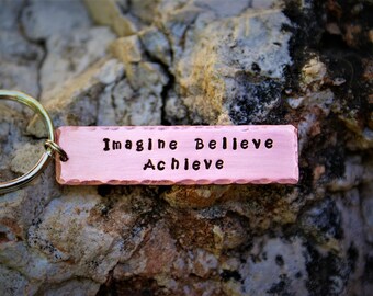 May include: A copper-colored rectangular keychain tag with the words "Imagine Believe Achieve" stamped in black. The tag is attached to a gold-colored keyring. The background is a textured rock surface.