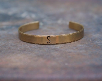 May include: A gold-toned metal bracelet with a hammered finish and the letter "S" engraved on the front.
