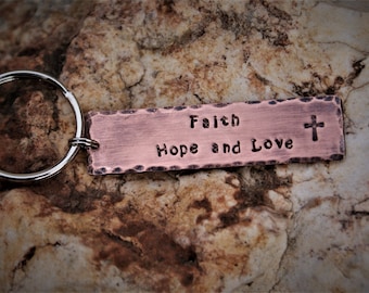 May include: A copper keychain with a rectangular tag that reads "Faith Hope and Love" with a small cross symbol at the end. The keychain is on a brown, rocky surface.