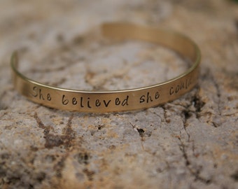 May include: A gold-toned metal bracelet with the inscription "She believed she could" engraved on the band.