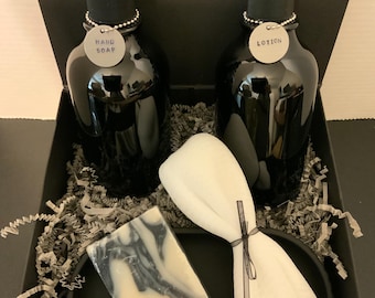 May include: A black gift set containing two black pump bottles labeled "HAND SOAP" and "LOTION", a bar of marbled soap, and a white washcloth tied with a black ribbon. The items are arranged in a black box with decorative paper.