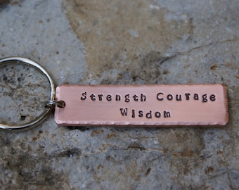 May include: A copper-colored rectangular keychain tag with the words "Strength Courage Wisdom" stamped into the metal. The tag is attached to a silver-colored key ring. The background is a textured, gray stone surface.
