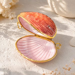 Könnte beinhalten: Eine geöffnete, aufklappbare muschelförmige Schatulle mit einem goldfarbenen Metallrand. Die Außenseite der Muschel hat eine texturierte, rot-orangefarbene Verlaufsoptik, während das Innere mit einer glatten, glänzenden, blassrosa Oberfläche ausgekleidet ist.
