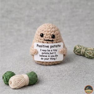 May include: A crocheted potato figure in tan yarn holds a sign that reads "Positive potato." The sign says, "I may be a tiny potato, but I believe in you. Go do your thing!" Two small yarn bundles are in the foreground.