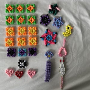 May include: A collection of colorful beaded crafts, including squares, stars, hearts, and a duck. The beads are in various colors such as green, orange, yellow, pink, blue, black, and purple. The crafts are arranged on a white surface.