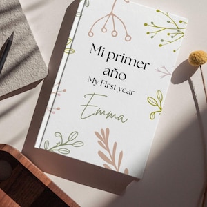Könnte beinhalten: Ein weißes Notizbuch mit dem Text "Mi primer año My First year Emma" in schwarzer und grüner Schrift. Das Cover zeigt botanische Illustrationen in gedämpften Rosa- und Grüntönen. Ein Bleistift und ein Notizblock sind im Hintergrund zu sehen.