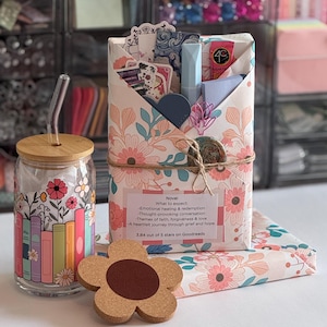 May include: A glass tumbler with a bamboo lid and glass straw featuring a colorful book and flower print, a flower-shaped cork coaster, and a rectangular gift package wrapped in floral paper with a label detailing book themes and ratings.