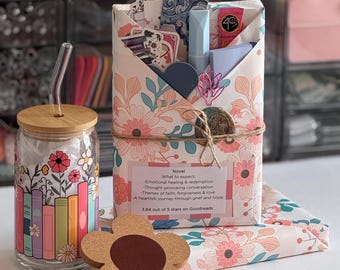 May include: A glass tumbler with a bamboo lid and glass straw featuring a colorful book and flower print, a flower-shaped cork coaster, and a rectangular gift package wrapped in floral paper with a label detailing book themes and ratings.