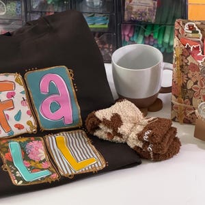 May include: A dark brown sweatshirt with the word FALL printed in large, colorful, patterned blocks. Beside it sits a white and brown ceramic mug, a pair of fuzzy brown and cream socks, a floral-patterned book sleeve, and a small glass candle.