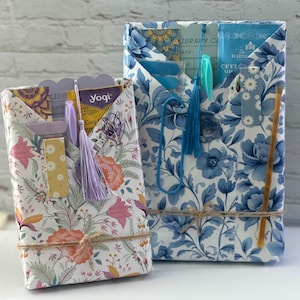 May include: Two gift-wrapped packages featuring floral patterns. The smaller package has a white background with pink, orange, and yellow flowers, a lavender tassel, and a Yogi tea bag. The larger package has a blue floral print with a blue tassel.