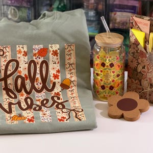 May include: A sage green sweatshirt featuring a graphic with vertical stripes, floral patterns, and the text "fall vibes" in brown script. Beside it sits a glass tumbler with a bamboo lid, a flower-shaped cork coaster, and a floral patterned stationery set.