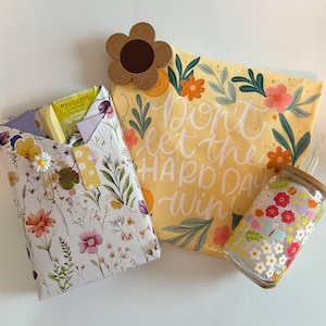 May include: A gift set featuring a floral-patterned paper envelope containing tea, a yellow fabric pouch with the text "Don't let the HARD DAY win!" surrounded by floral illustrations, a brown flower-shaped coaster, and a glass tumbler with a floral print.