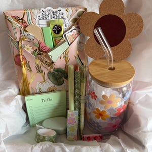 May include: A gift set containing a floral patterned envelope, a glass tumbler with a bamboo lid, a cork flower topper, a clear glass straw, a green To Do notepad, three pens, a small green container, and two rolls of decorative washi tape.