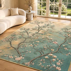 May include: A rectangular area rug featuring a teal and beige background with a sprawling tree branch pattern. The design includes delicate light pink blossoms and green leaves extending across the surface, finished with a thin gold-toned border.
